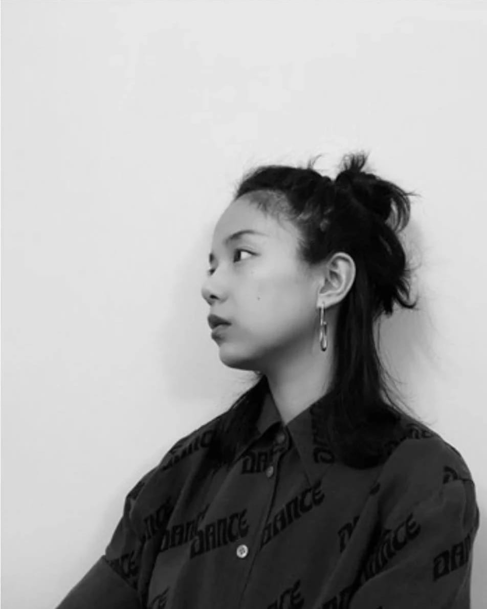 A black and white image of an Asian woman wearing a black blouse agains the backdrop of a white/gray wall, staring off to the left. 