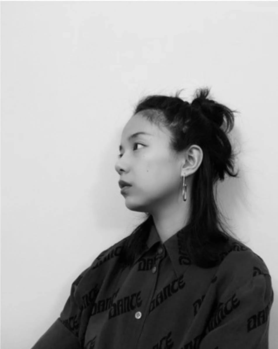 A black and white image of an Asian woman wearing a black blouse agains the backdrop of a white/gray wall, staring off to the left. 