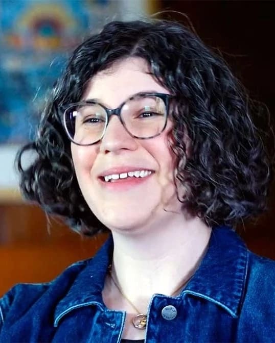 A video still of a person wearing a blue jean jacket with black curly hair and glasses who is smiling at someone offscreen.