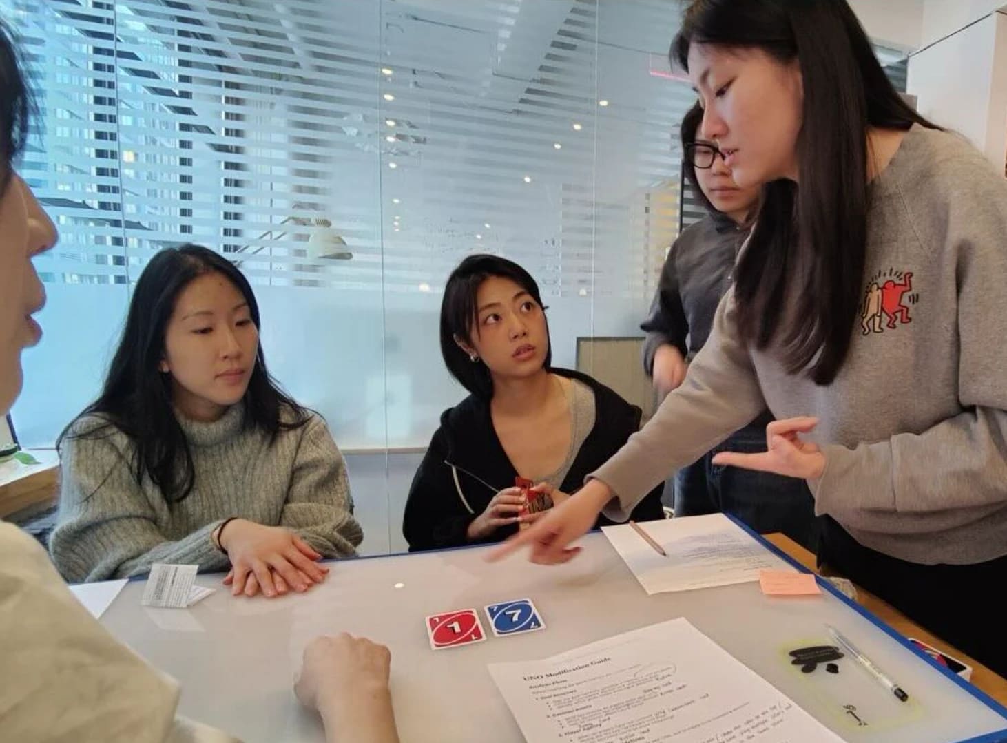 A group of students gathered around a table and discussing the card game Uno.