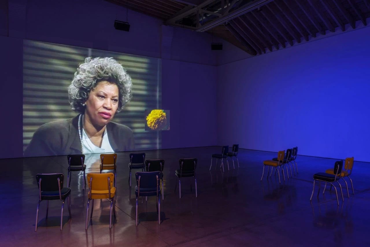 Picture of a museum installation of a large projected screen with a woman and a yellow flower. Several chairs are organized in front of the projection.