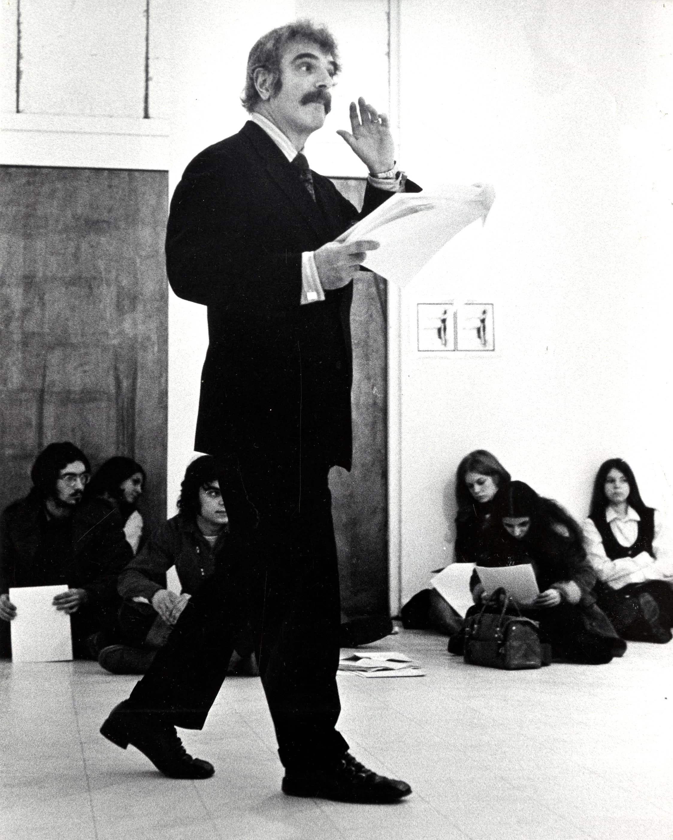 An archival photograph of a mustachioed man in a suit, standing and holding papers and lecturing to a class of students, who are seated on the floor behind him