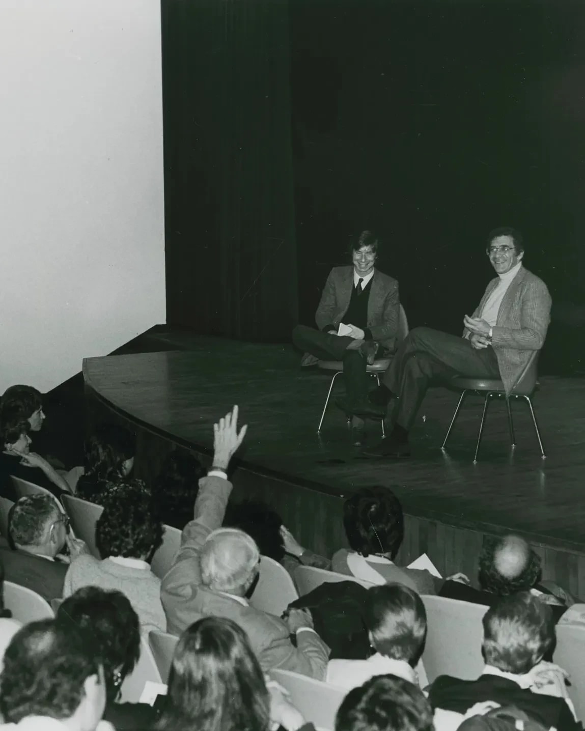 A black and white photo of two people sit on stage in front of a crowd.