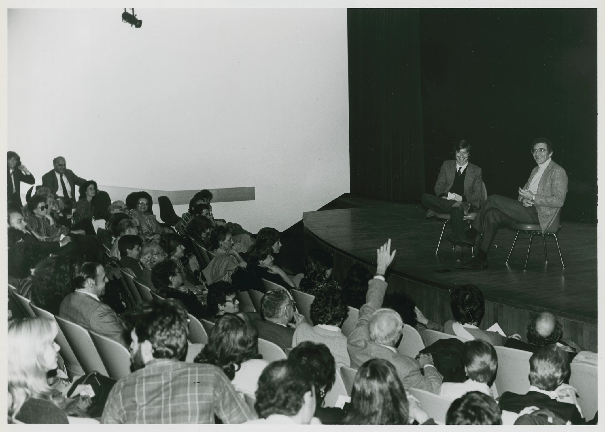A black-and-white photo of two people sitting on an empty stage in front of a crowd. A person in the crowd has raised their hand to ask a question