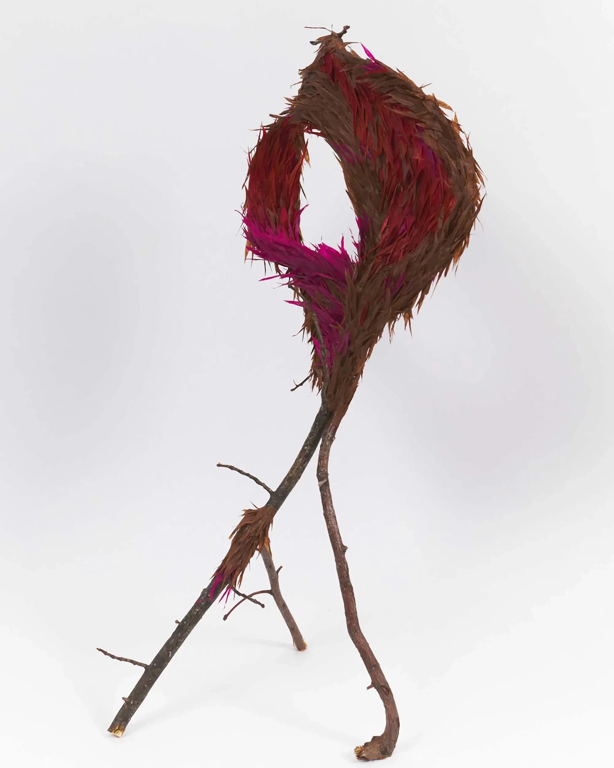 A whimsical sculpture of branches forms a loop at the top, adorned with vibrant magenta and brown feathers, set against a plain white background.