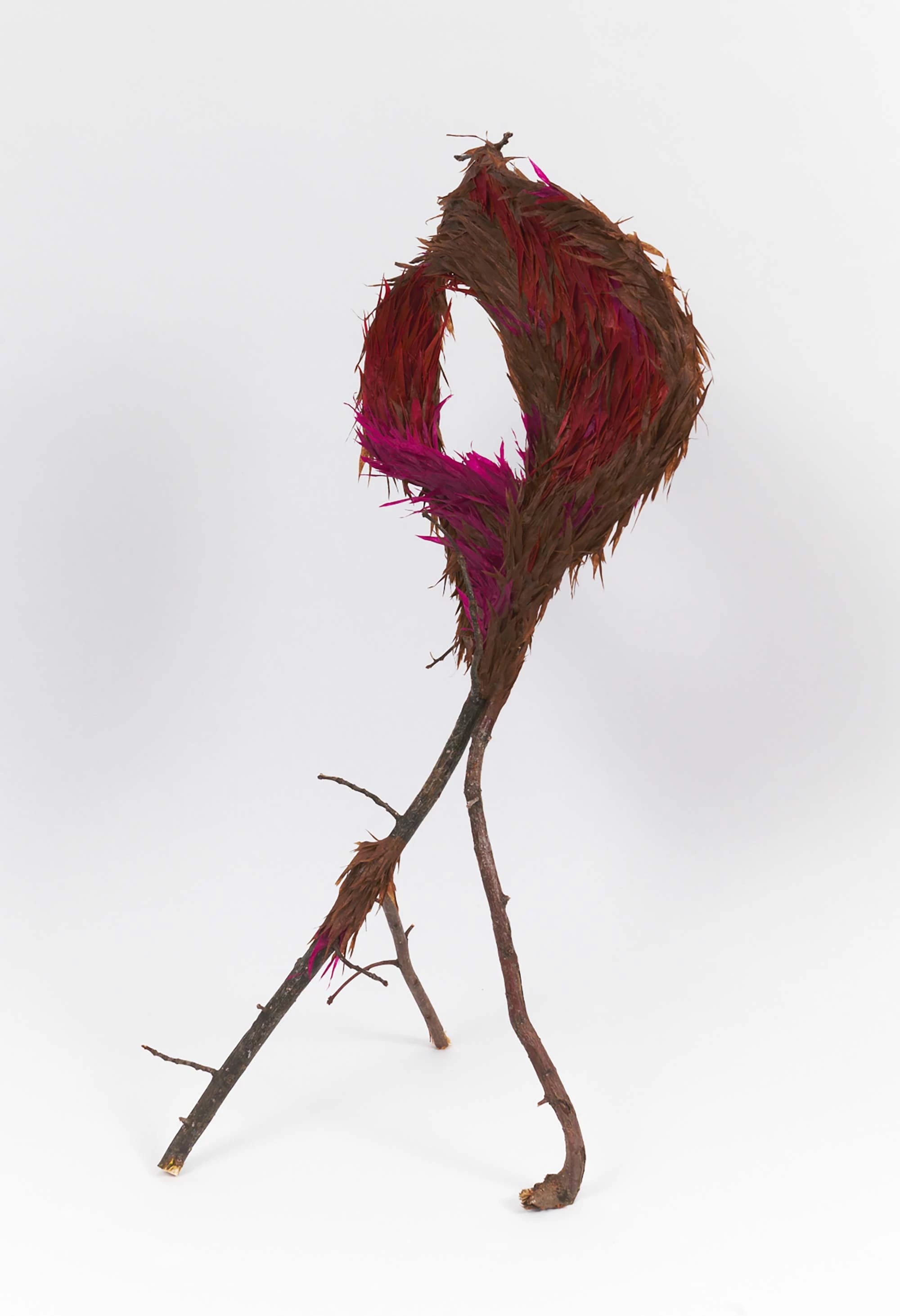 A whimsical sculpture of branches forms a loop at the top, adorned with vibrant magenta and brown feathers, set against a plain white background.