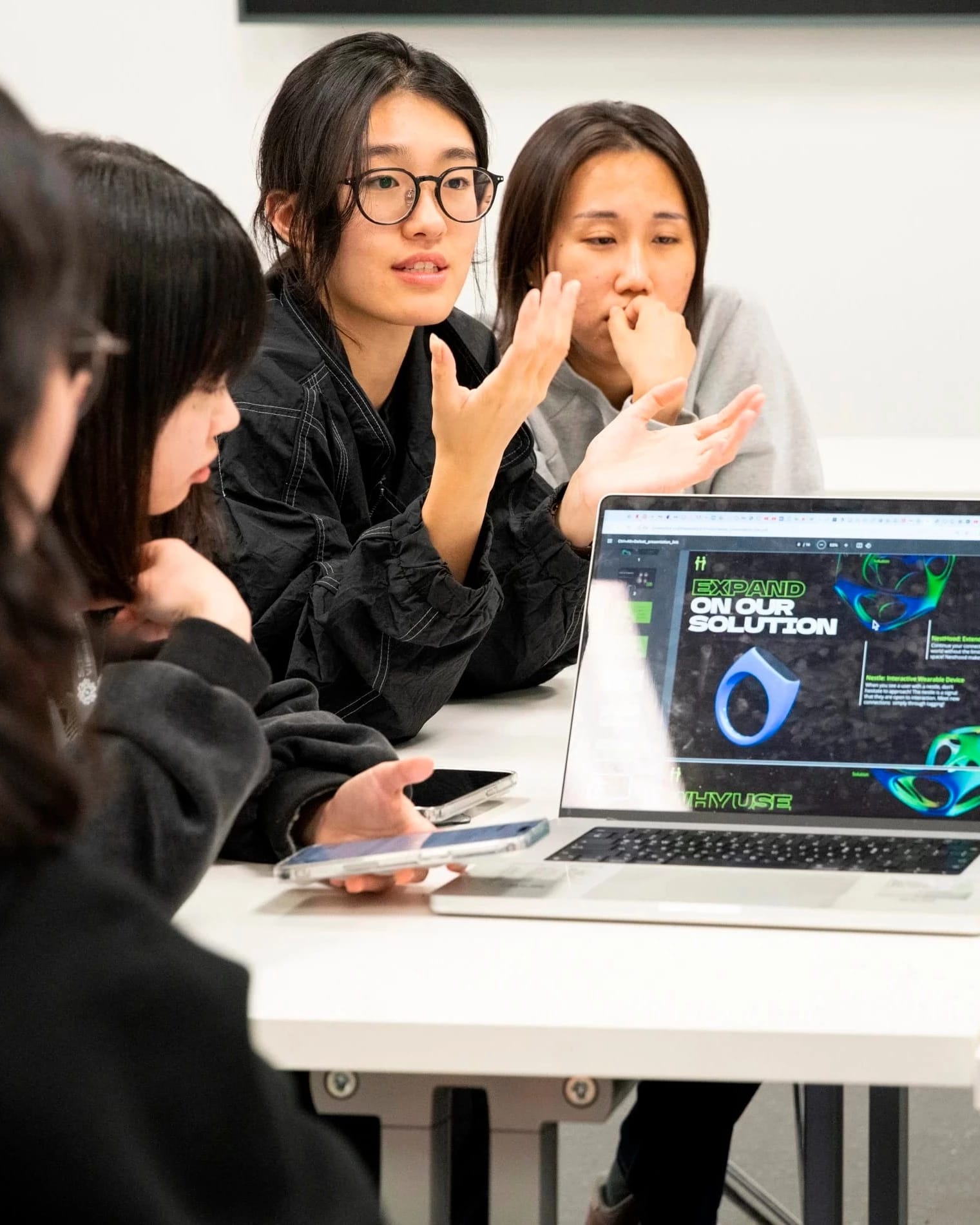 Four students sit at a table and present their work behind a laptop screen showing their presentation.