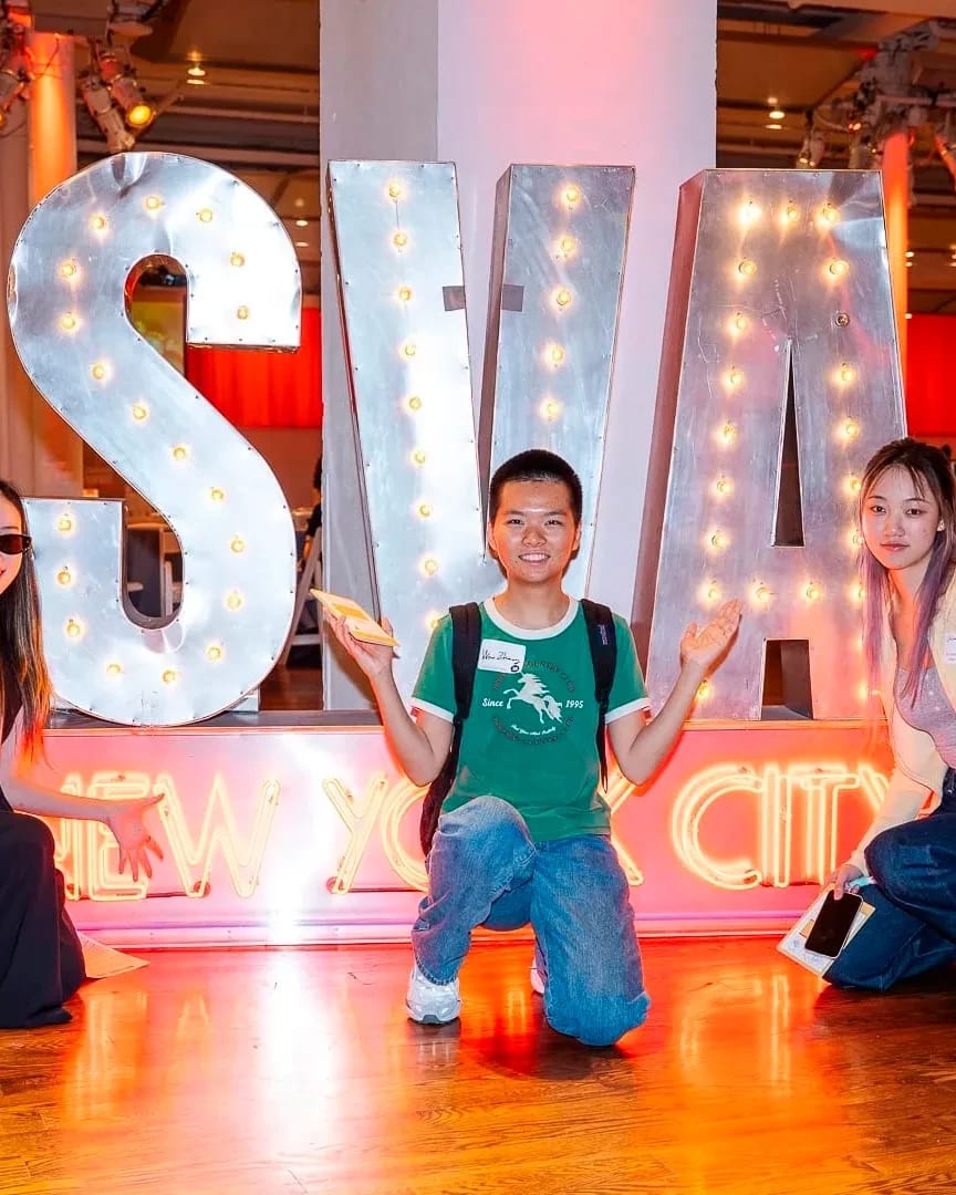 Three students pose in front of a large illuminated “SVA New York City” sign at an indoor welcome event.
