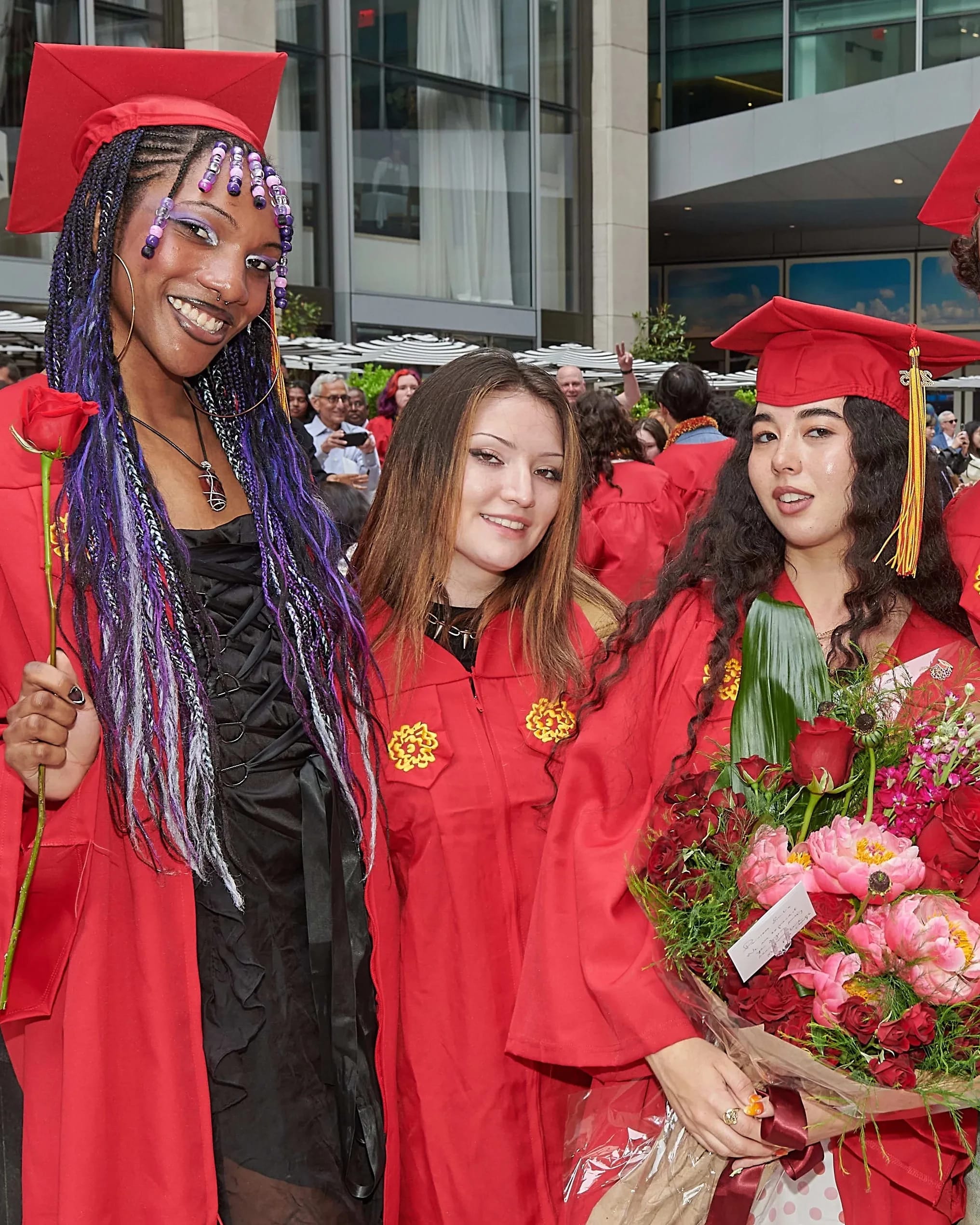 Three graduates wearing red commencement regalia pose for a photo; the right-most person is holding a large bouquet of pink and red flowers.