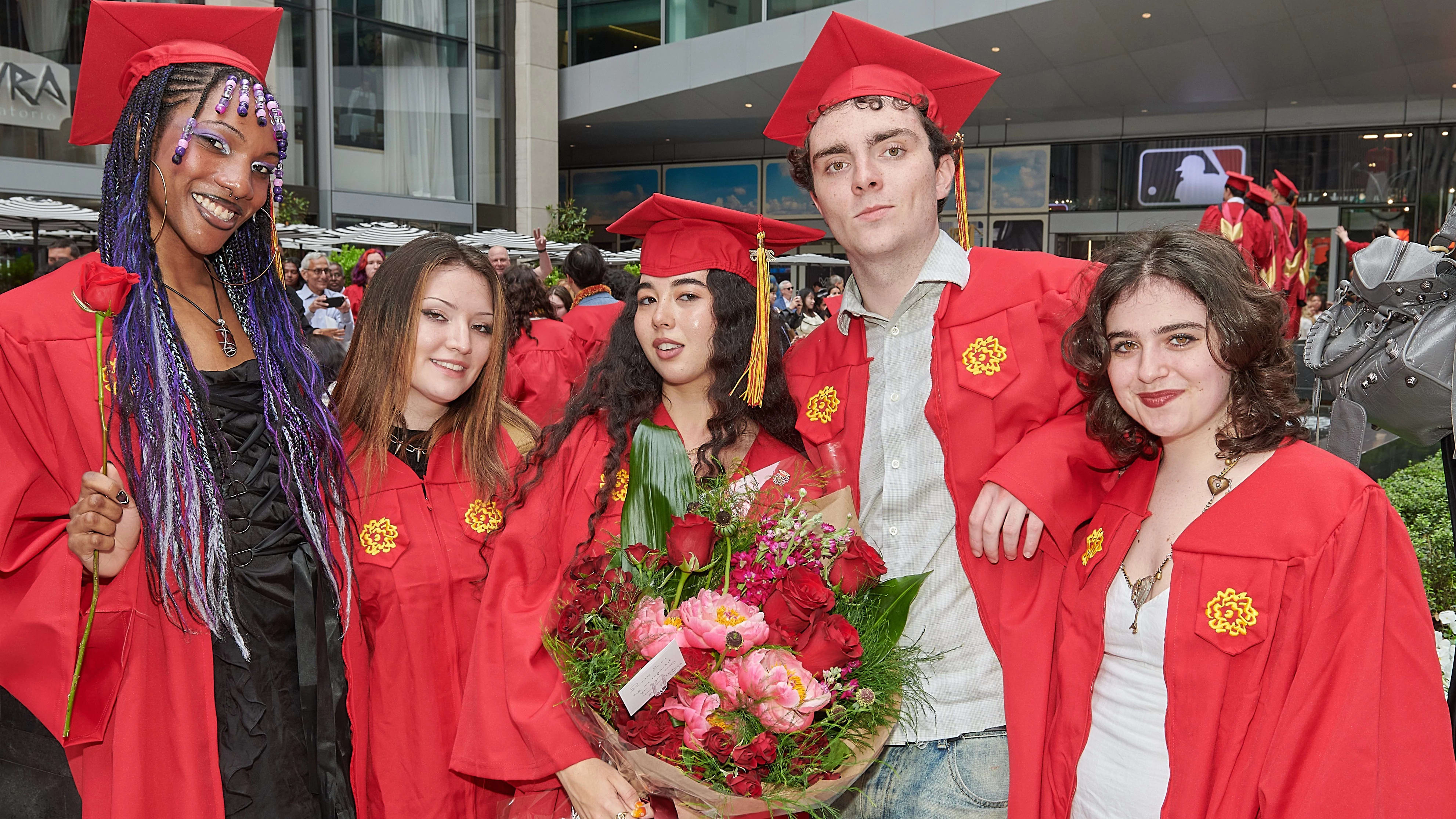 Five graduates wearing red commencement regalia pose for a photo; the middle person is holding a large bouquet of pink and red flowers.