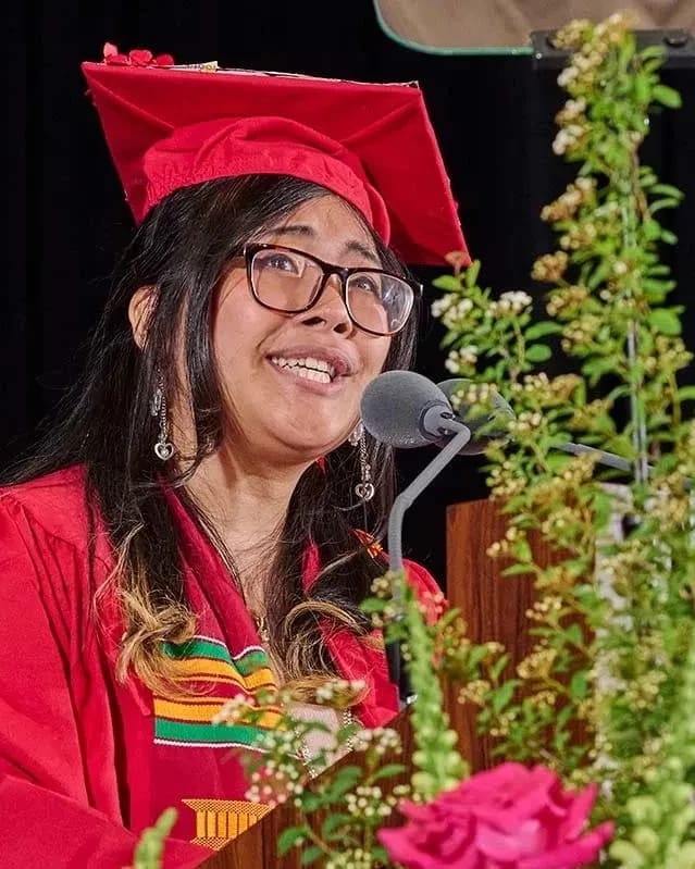 A graduate in a red cap and gown speaks passionately at a podium adorned with greenery and flowers. The background is a dark curtain, conveying a celebratory tone.