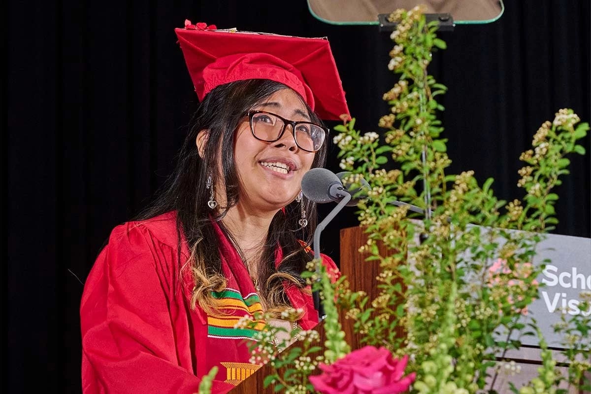 A graduate in a red cap and gown speaks passionately at a podium adorned with greenery and flowers. The background is a dark curtain, conveying a celebratory tone.