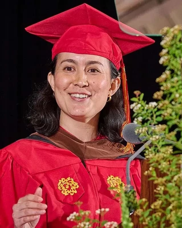 Graduate woman in red cap and gown delivers a speech at a lectern with greenery. She smiles warmly, exuding a sense of achievement and joy.