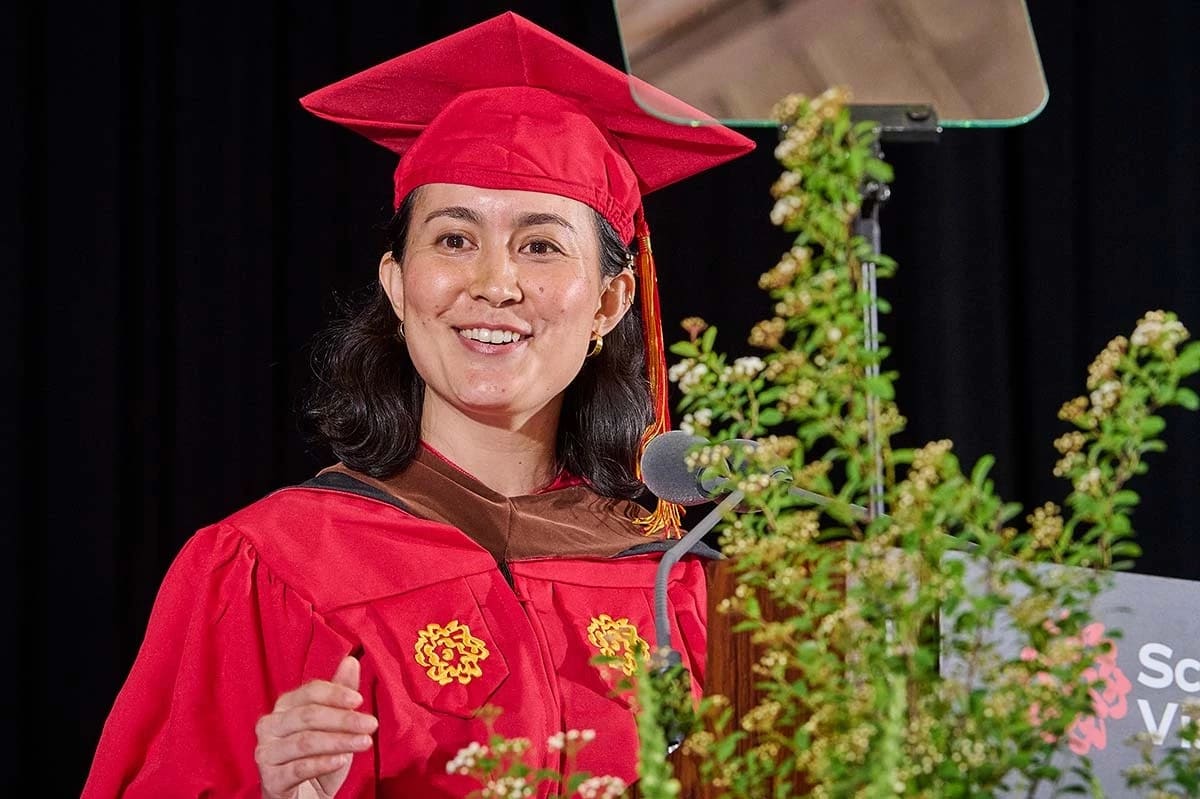 Graduate woman in red cap and gown delivers a speech at a lectern with greenery. She smiles warmly, exuding a sense of achievement and joy.