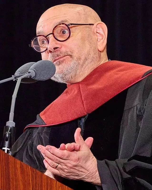 A bald man with glasses and a trimmed beard speaks at a podium, wearing a black academic robe with a red hood. Expression is thoughtful and engaging.