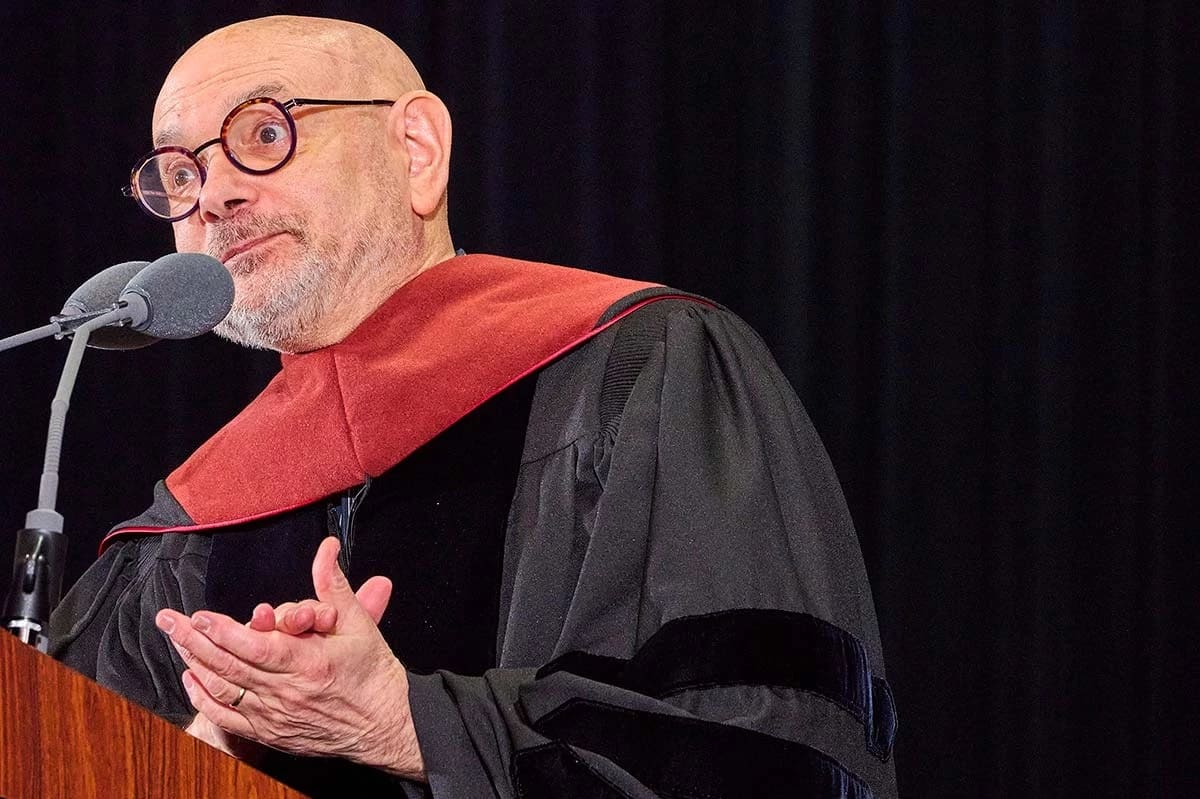 A bald man with glasses and a trimmed beard speaks at a podium, wearing a black academic robe with a red hood. Expression is thoughtful and engaging.