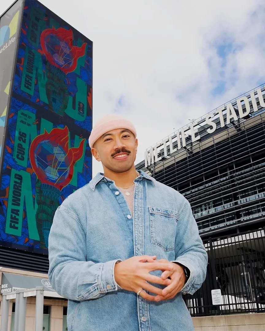 A man stands in a blue denim jacket and a pink beanie with his hands clasped in front of him. He's smiling and standing in front of the MetLife Stadium. A large vertical billboard displays Rich Tu's new poster.