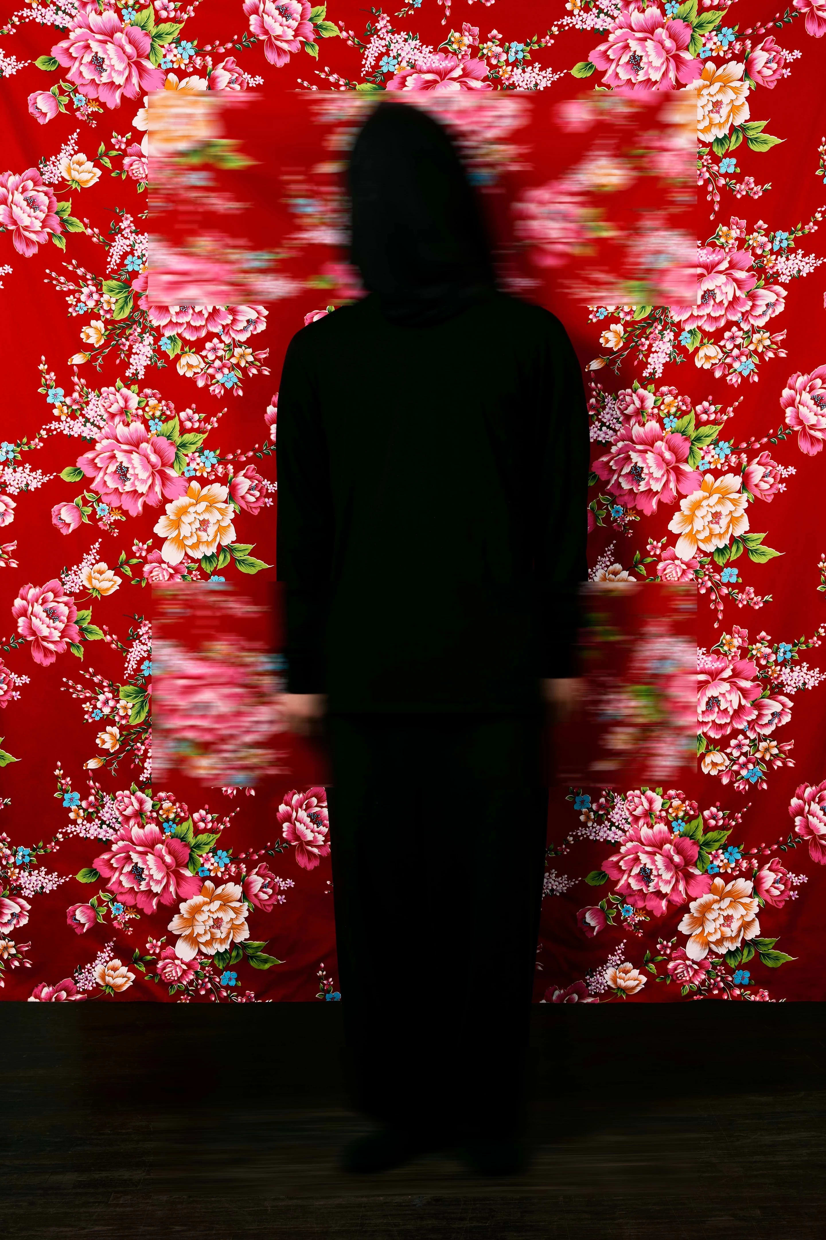 A black silhouette stands in front of a bright red floral wallpaper with two rectangular patches blurred.