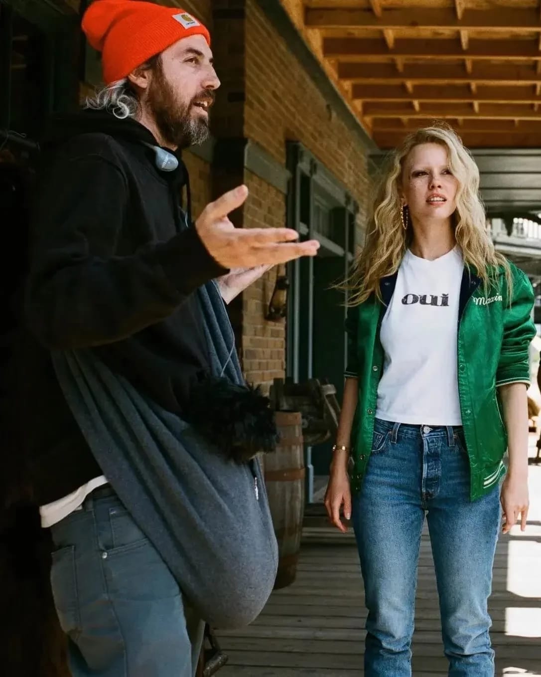 A man with long hair wearing a orange winter cap stands with his arms open, gesturing, with a blonde woman to his right wearing a bright green jacket and blue jeans. 
