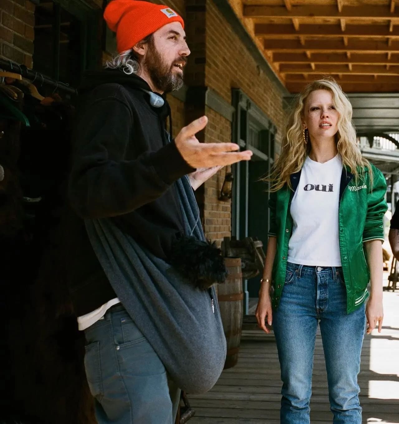 A man with long hair wearing a orange winter cap stands with his arms open, gesturing, with a blonde woman to his right wearing a bright green jacket and blue jeans. 