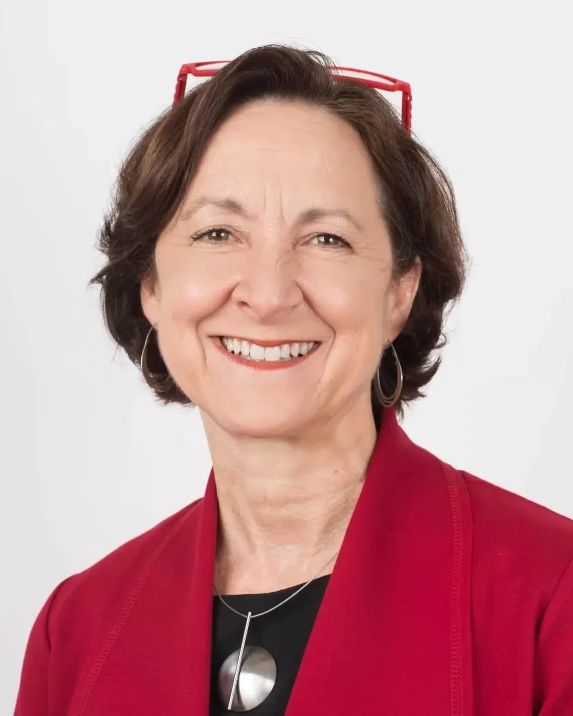 A woman with brown hair, red glasses sitting atop of her head wearing a red jacket, a black shirt, and silver medallion necklace looking at the camera and smiling. 