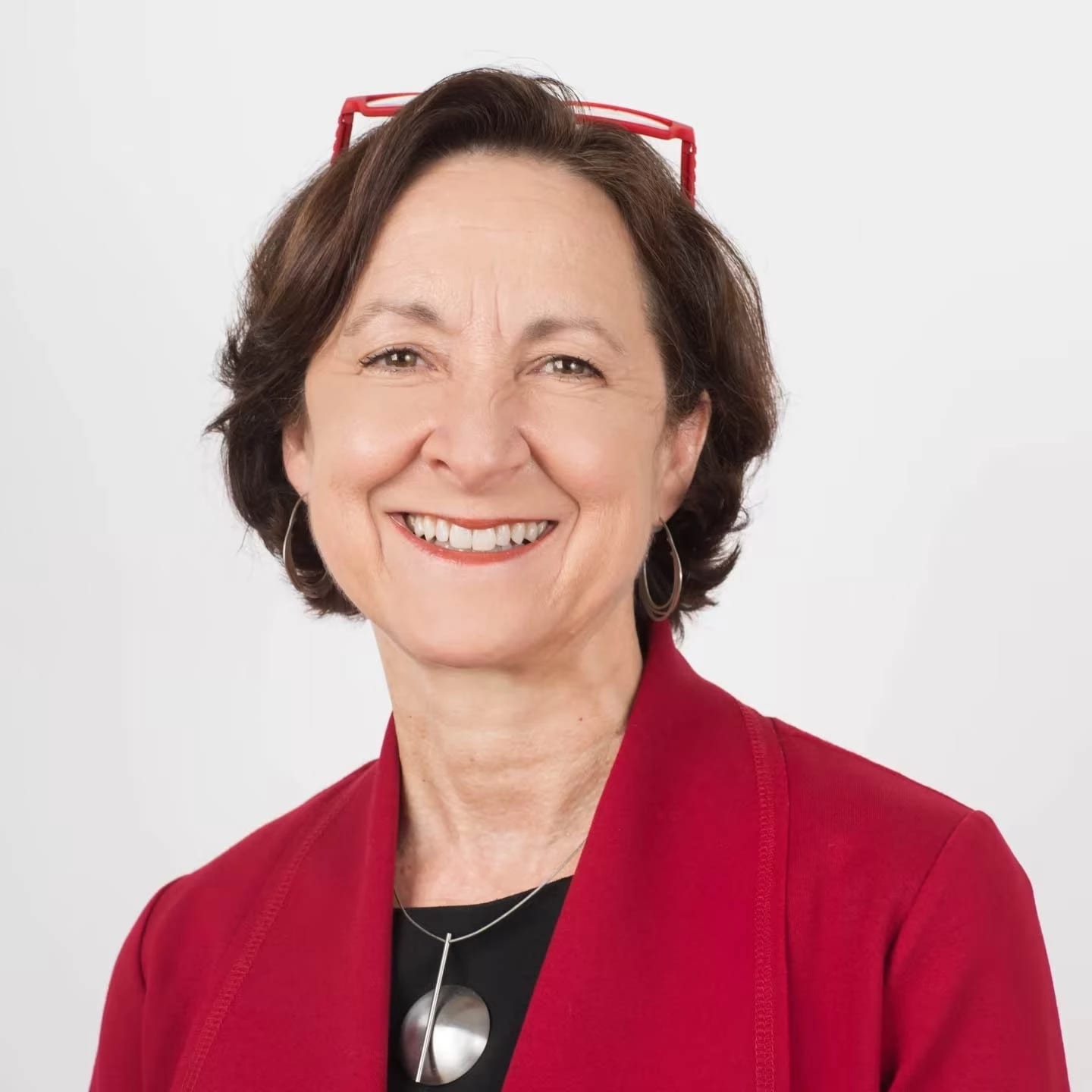 A woman with brown hair, red glasses sitting atop of her head wearing a red jacket, a black shirt, and silver medallion necklace looking at the camera and smiling. 
