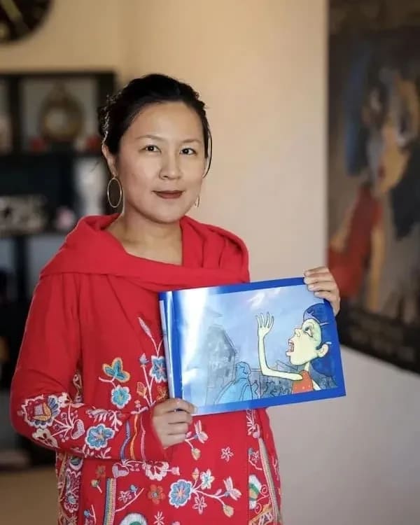 A woman in a red embroidered garment holds a children's book with an illustration of a shouting child. She stands indoors, conveying warmth and creativity.