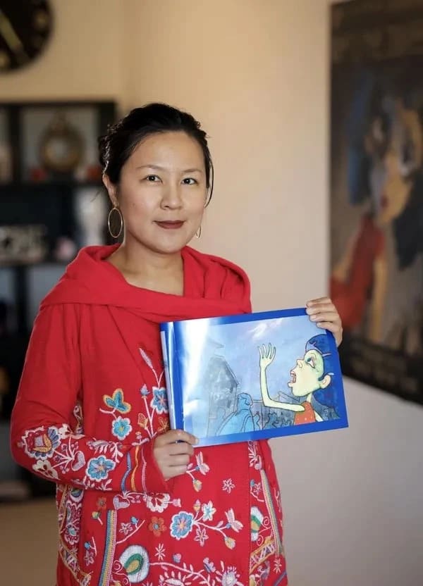 A woman in a red embroidered garment holds a children's book with an illustration of a shouting child. She stands indoors, conveying warmth and creativity.
