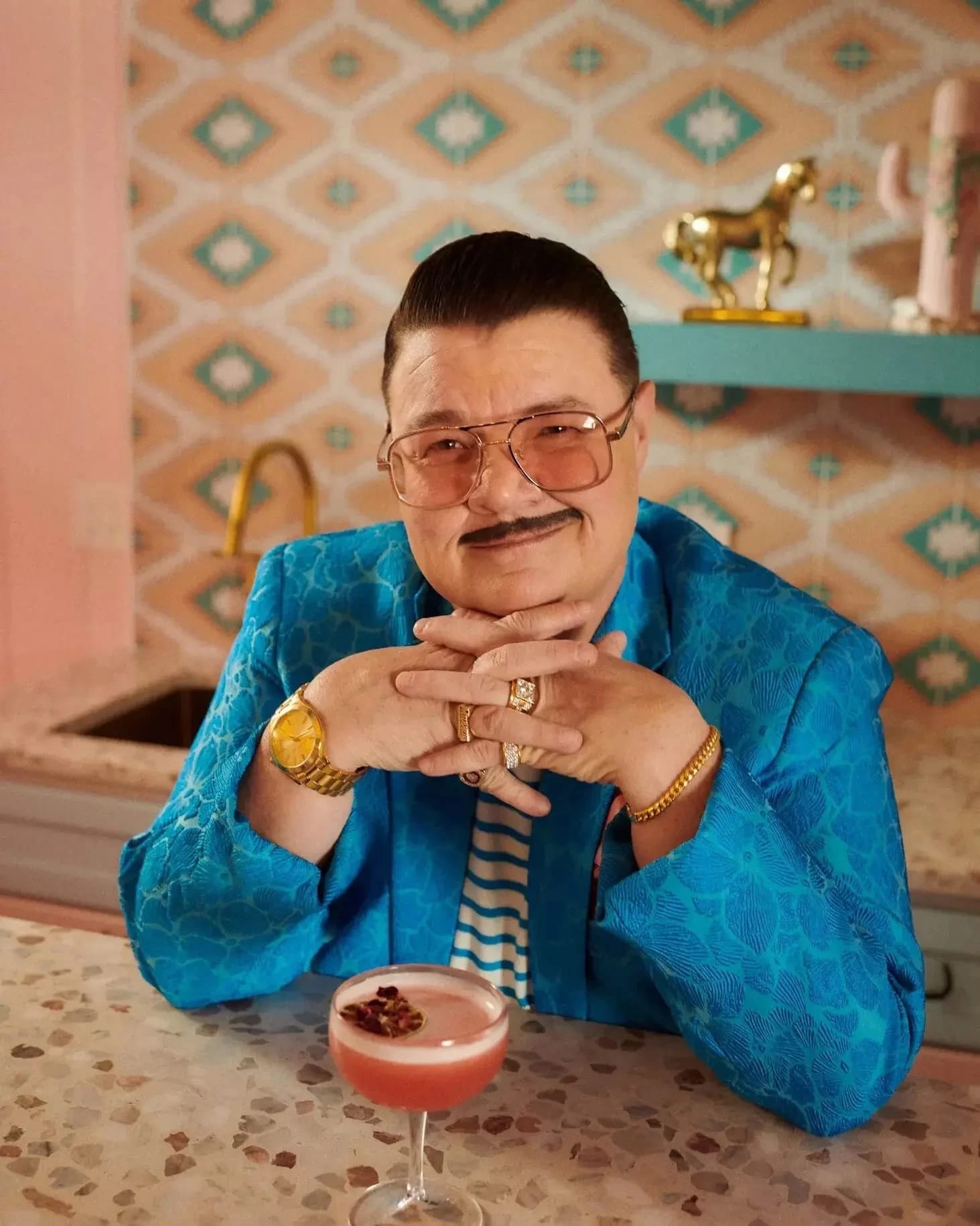 A man with pencil thin mustache and sunglasses, wearing a electric blue suit smiles at the camera.