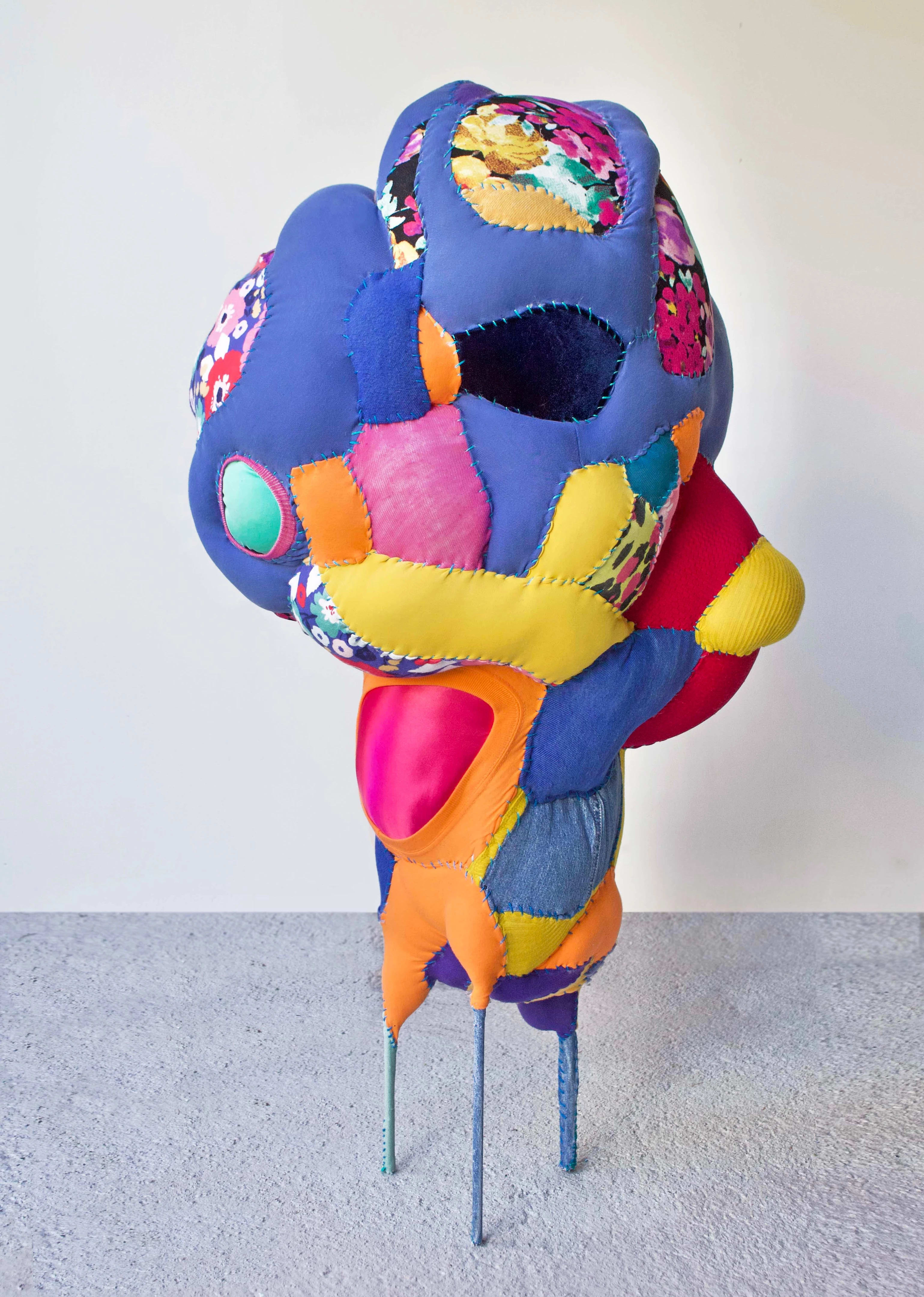 Recycled textile sculpture made of thread, batting and found steel. It is a bunch of different colored fabrics sewn together to create a top heavy blob that is sitting on a stool.