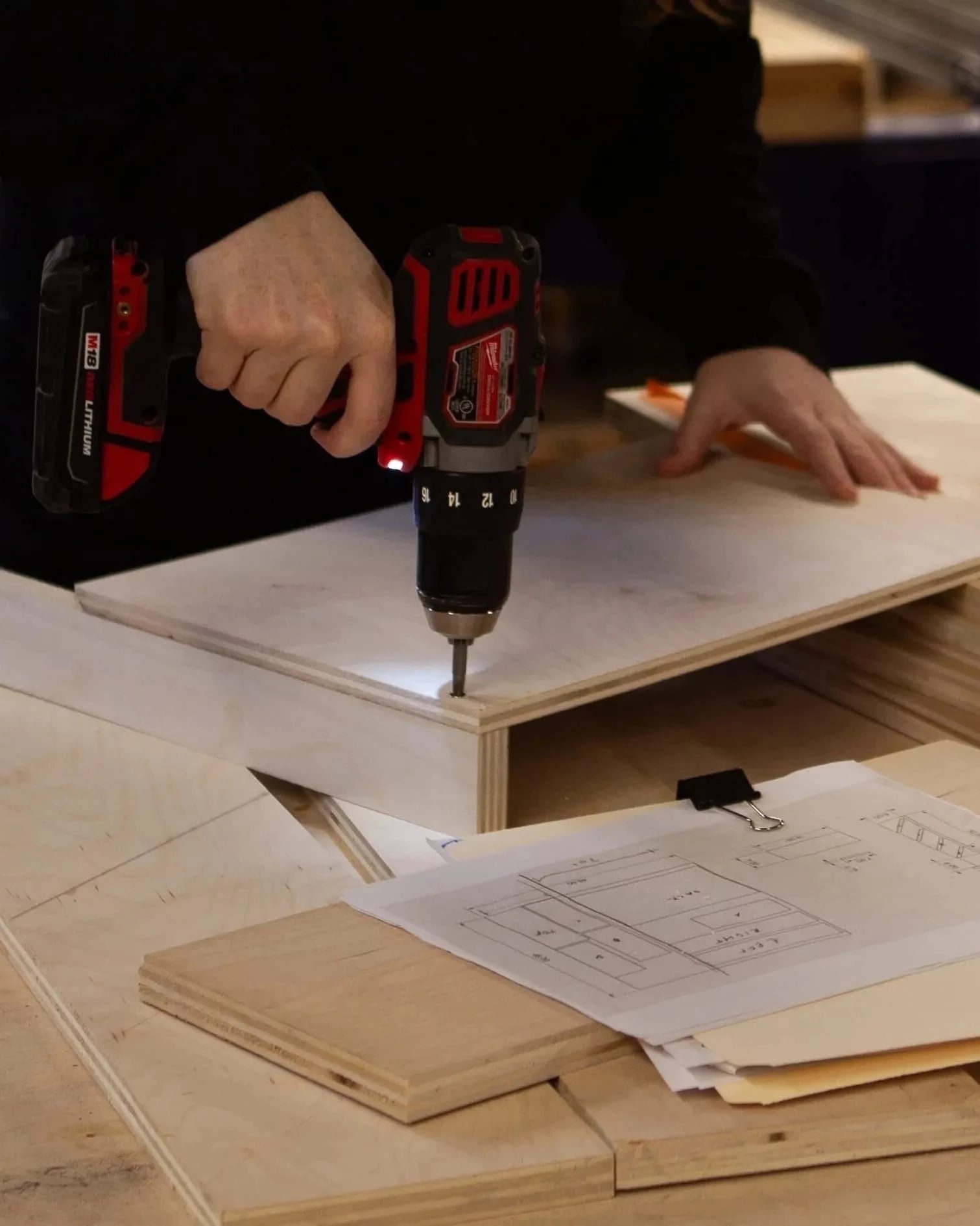Hand using a drill on a piece of wood. Measuring tape, ruler, and other tools on the table.