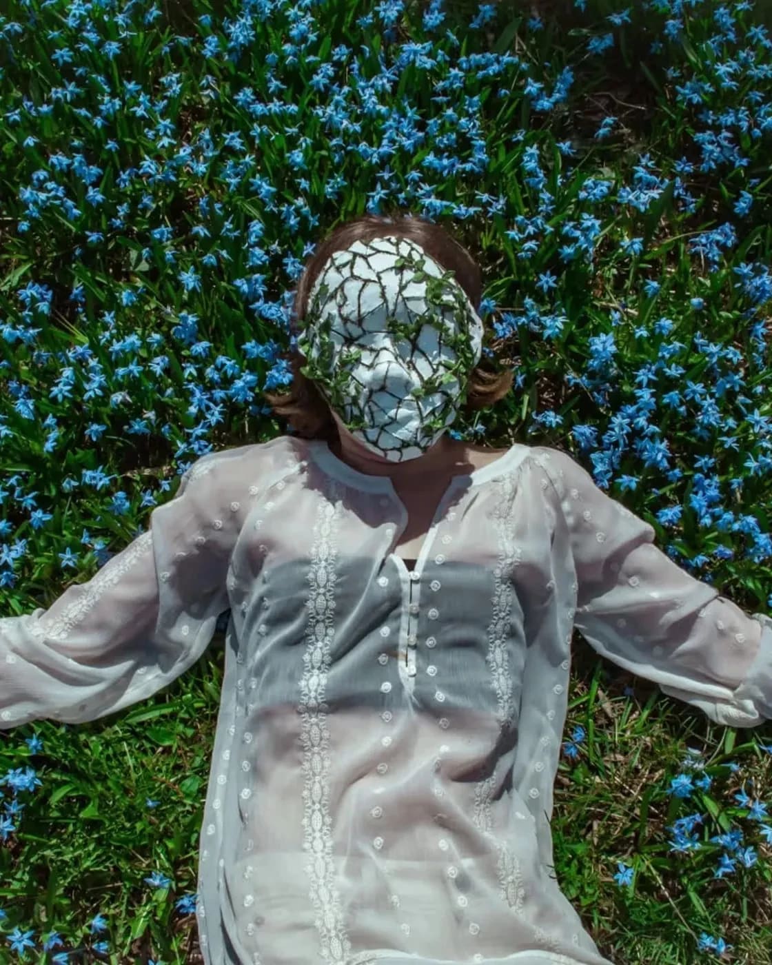 a pale person in a sheer white shirt lies in a field of flowers. There's a mask on their face that's cracked and has leaves growing out of it. 