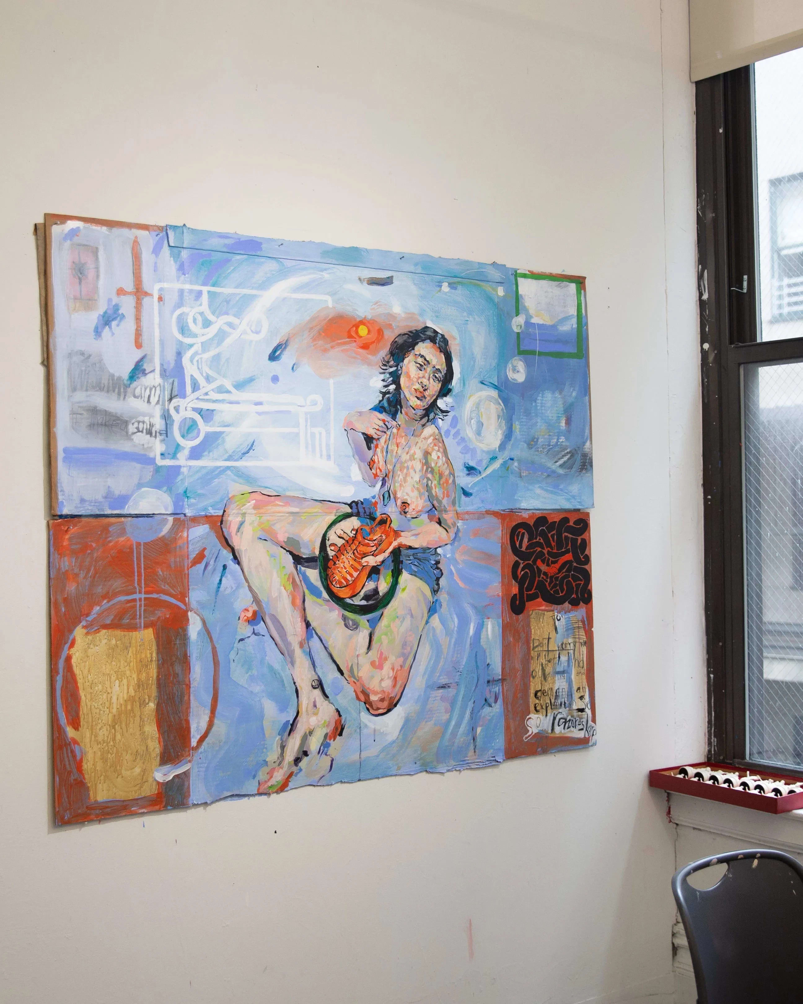 Windowed artist studio in NYC with paintings on the wall and a desk.